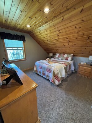 A bedroom with a sloped ceiling, a bed with a quilt, a desk with a lamp, and a window with blinds.