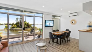 Habitación moderna con un amplio ventanal corredero que da a un balcón con vistas a un lago. Adentro, hay una mesa de comedor de madera con sillas, un televisor montado en la pared y una pequeña mesa redonda.