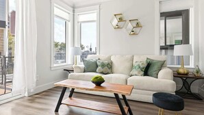 Una sala de estar moderna con un sofá, una mesa de centro de madera y un reposapiés negro. Se observan dos decoraciones hexagonales en la pared y una lámpara de pie.
