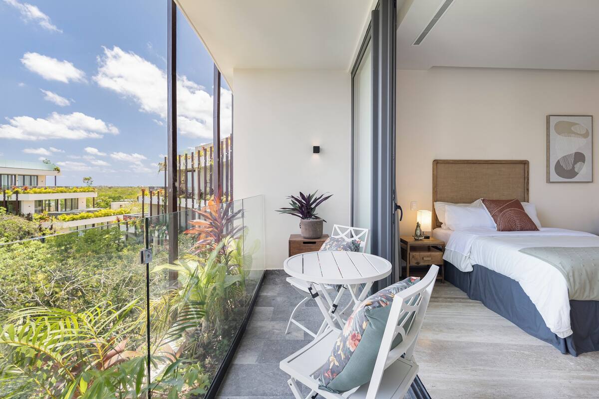 A modern hotel room with a balcony, a bed, a bedside table with a lamp, and a view of greenery and a building outside.