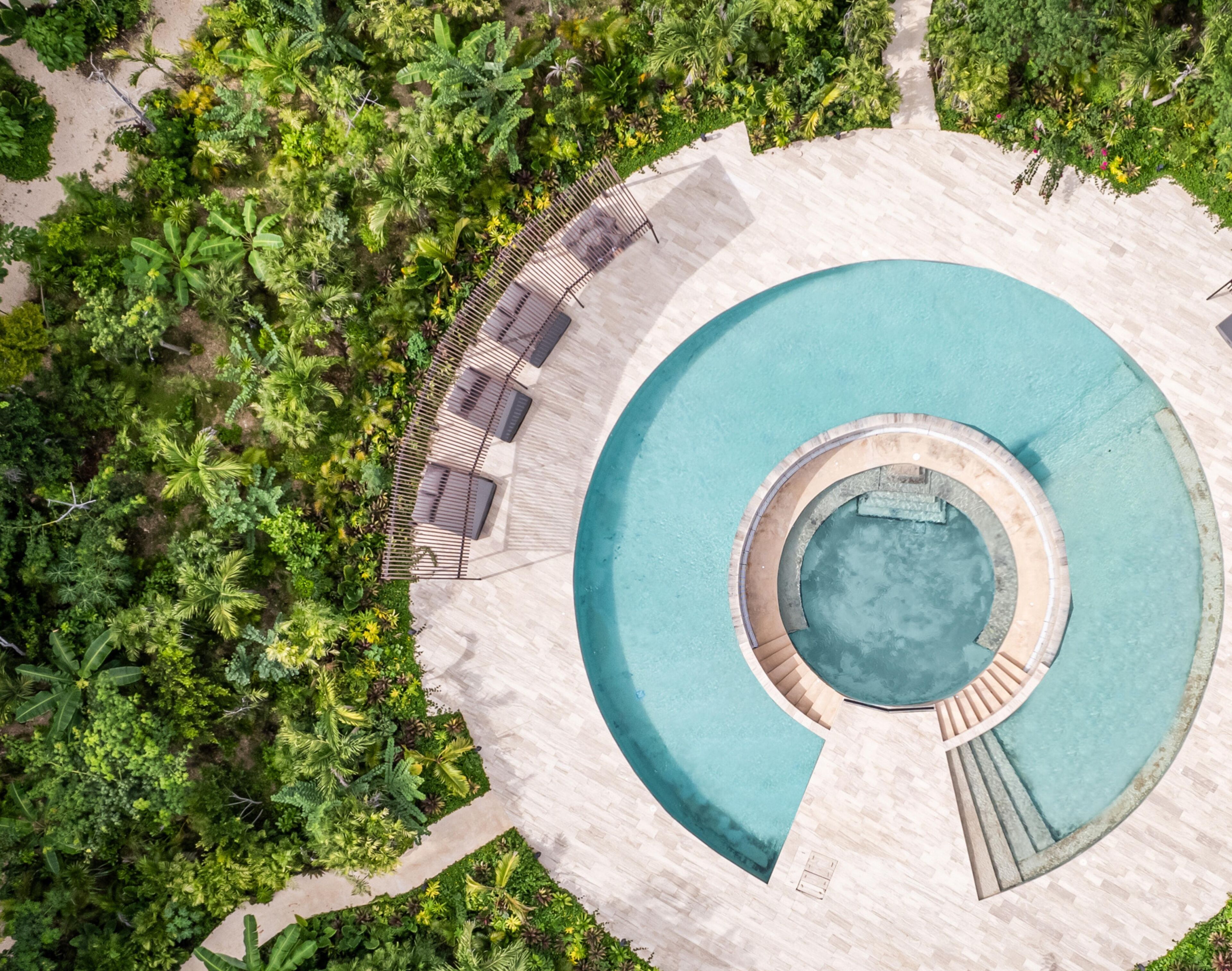An aerial view of a circular swimming pool with a central hot tub, surrounded by lush greenery and a tiled deck with outdoor seating.