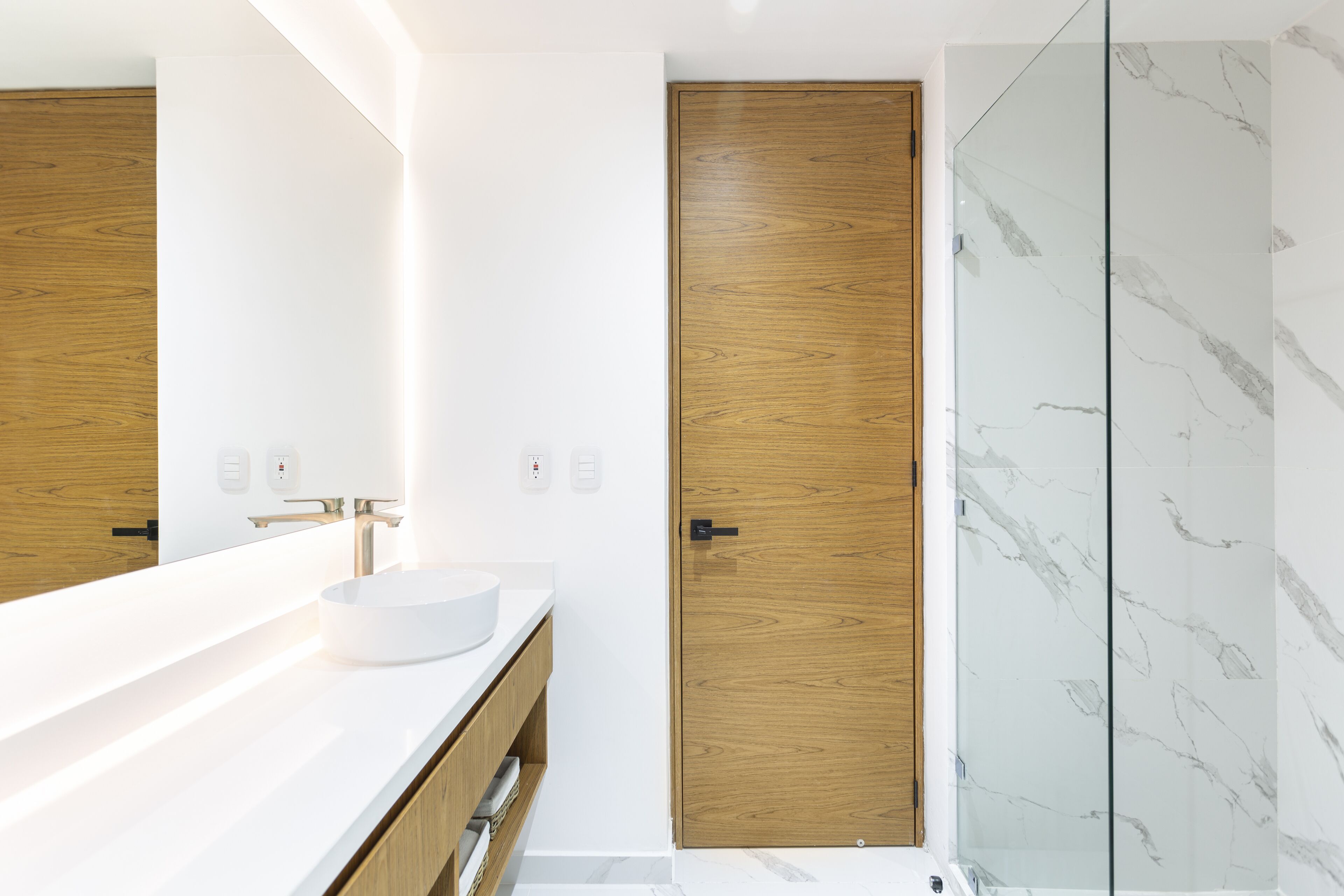 Un baño moderno con lavamanos blanco, tocador de madera y cabina de ducha de vidrio.