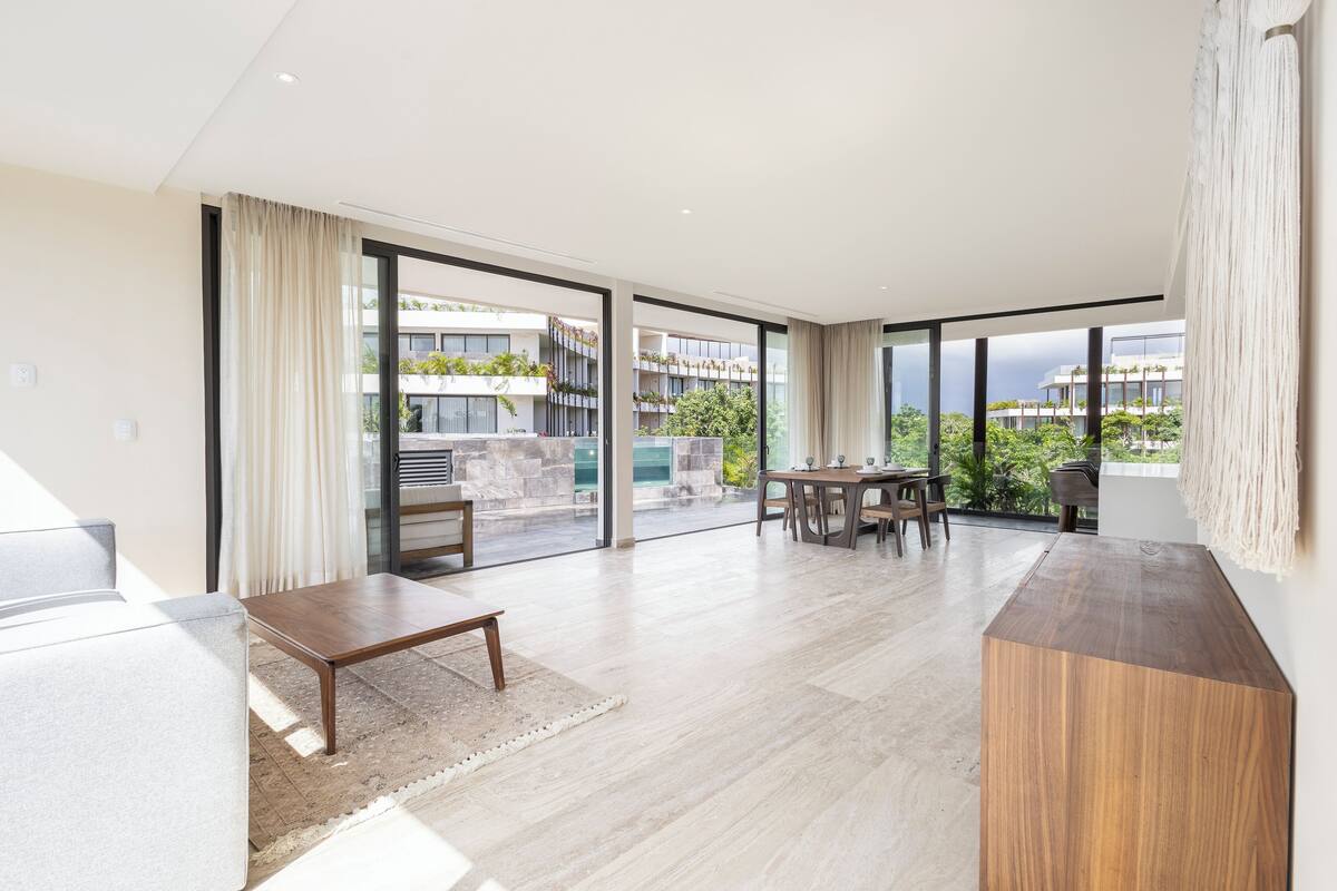 A spacious living area with a wooden coffee table, a sofa, and a dining table with chairs. Large windows offer a view of a building and greenery outside.