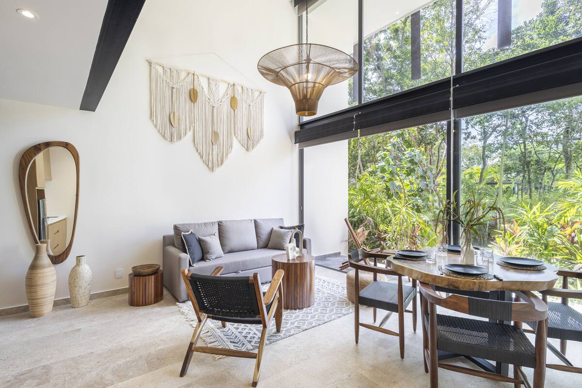 A modern living room with a gray sofa, wooden dining table, and a large window overlooking greenery.