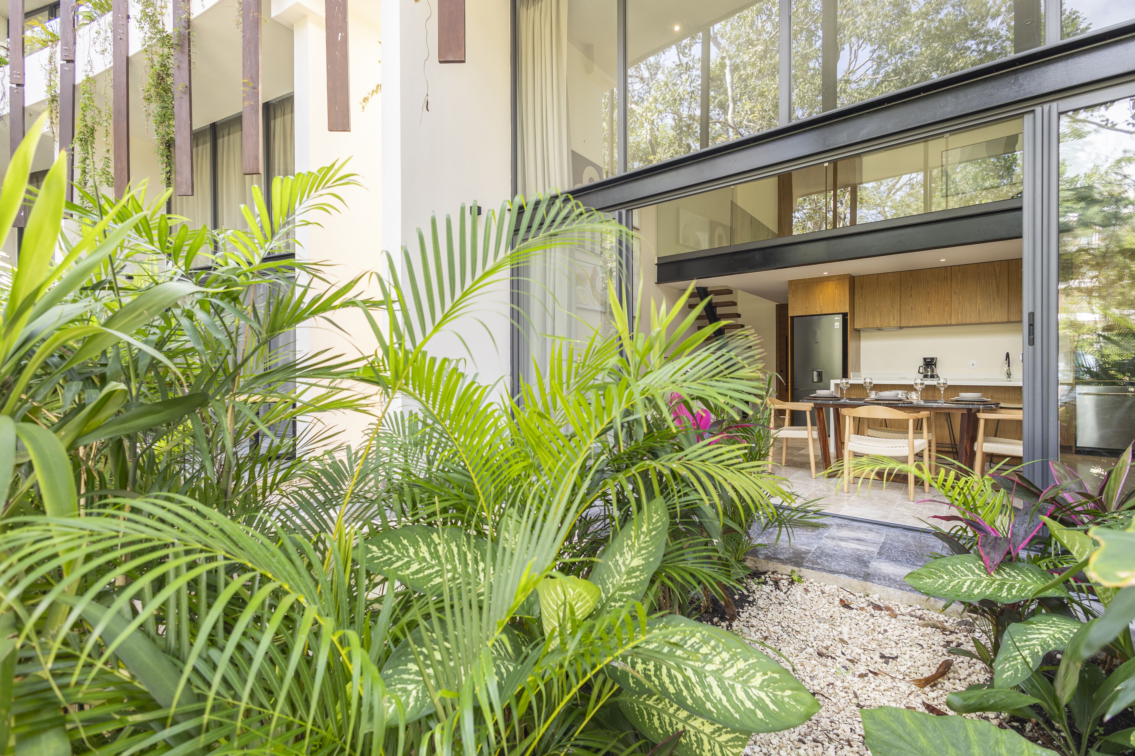 A modern house with a patio area featuring a dining table and chairs, surrounded by lush green plants and a gravel path.