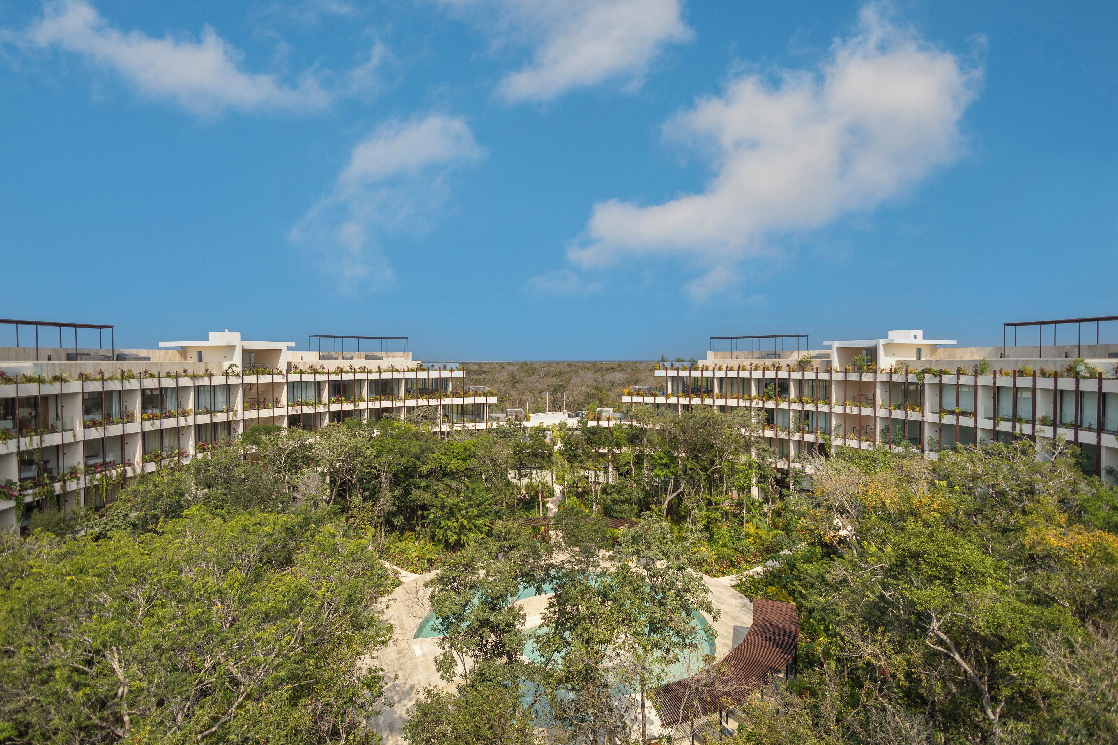 A modern hotel with a central pool surrounded by lush greenery, multiple floors with balconies, and a clear blue sky.