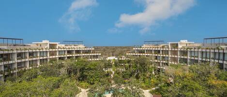 A modern hotel with a central pool surrounded by lush greenery, multiple floors with balconies, and a clear blue sky.