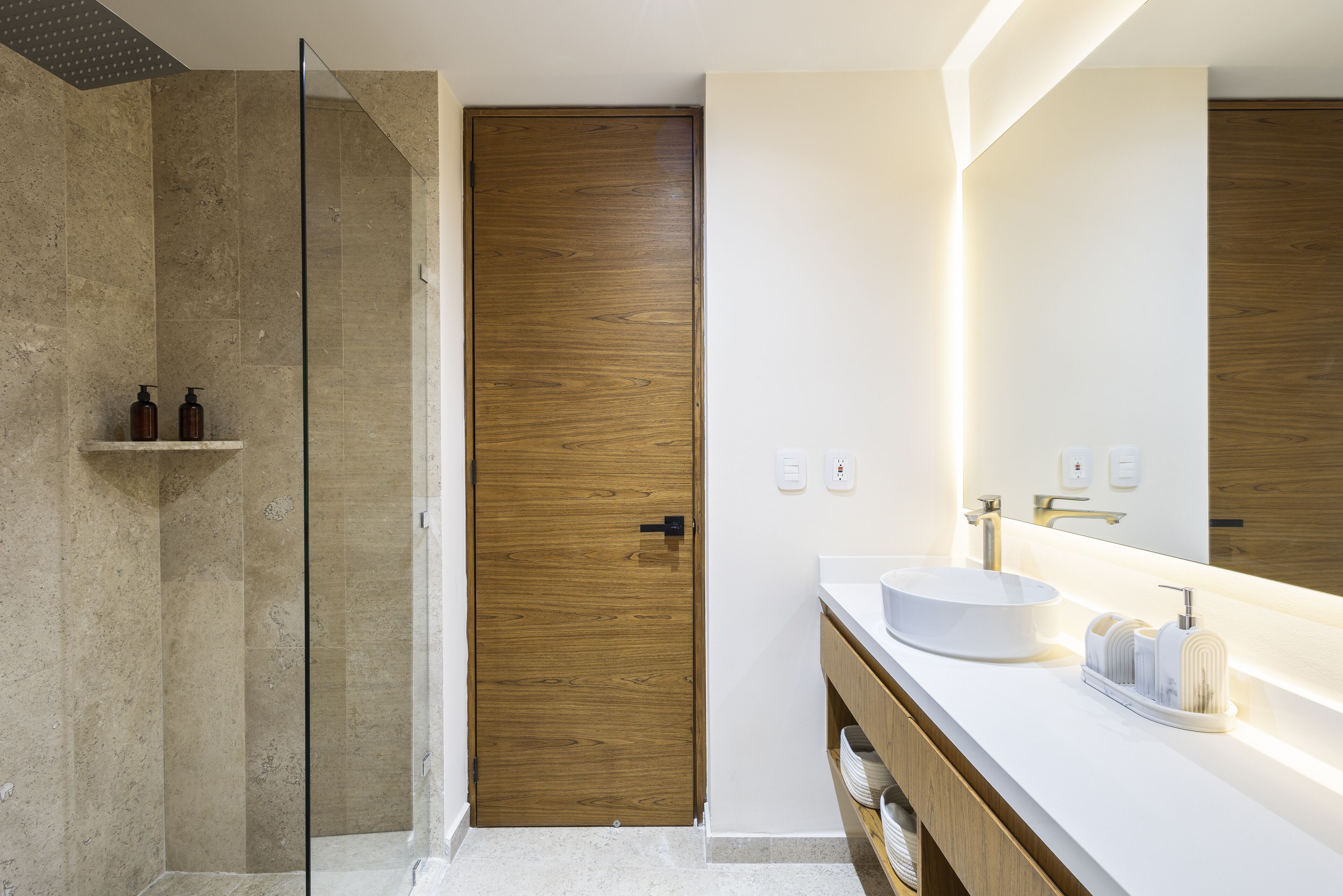 Un bagno moderno con porta in legno, cabina doccia in vetro, lavabo bianco e uno specchio grande.