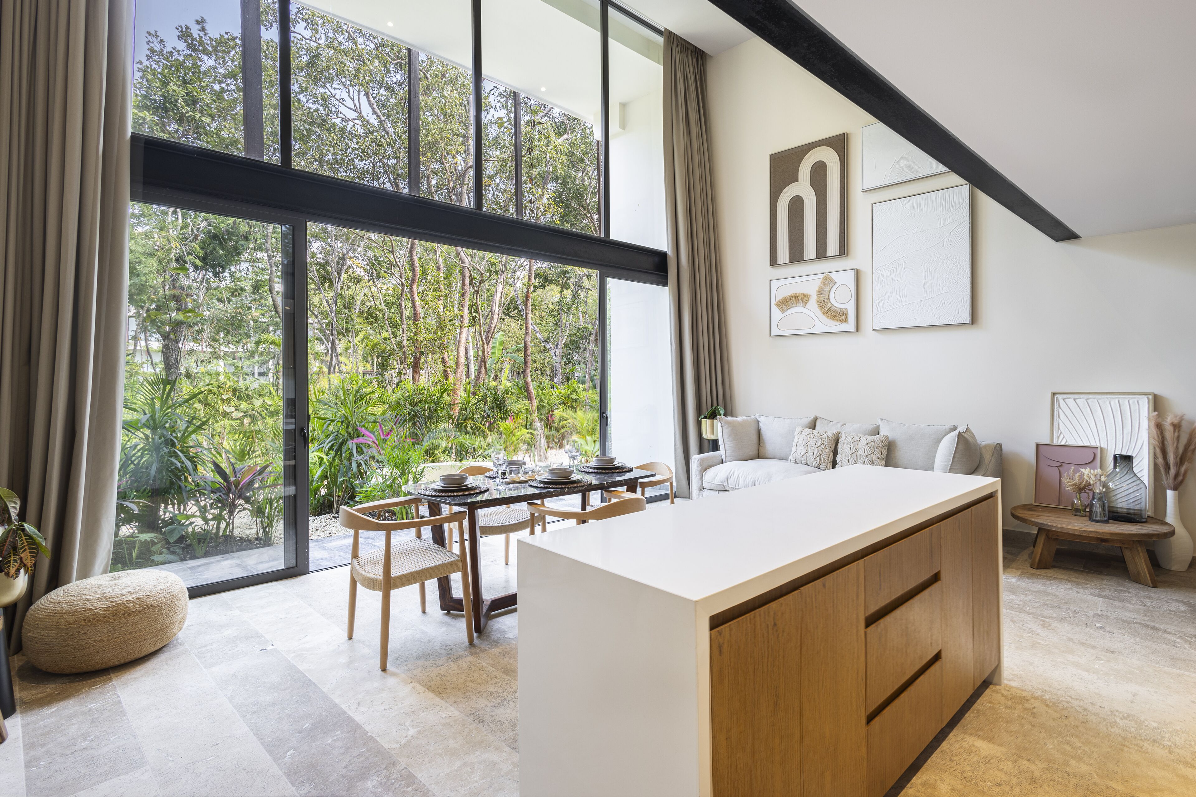 Une cuisine moderne dotée d’un grand îlot central, d’un coin repas avec une table et des chaises, et offrant une vue sur la verdure grâce à de grandes fenêtres.