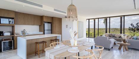 A modern kitchen with a central island, a dining area with a wooden table and chairs, and a living area with a sofa and a view of the outdoors.