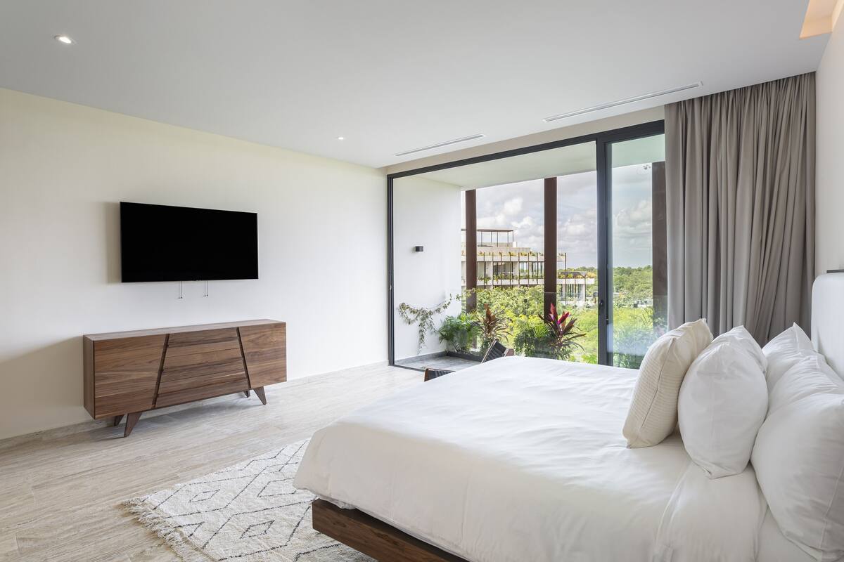 A modern bedroom with a large bed, a flat-screen TV mounted on the wall, and a view of greenery through a large window.