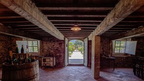 A rustic cellar with stone walls, wooden beams, and a brick floor. There are two windows with open shutters, a wooden barrel, and a small table with bottles.