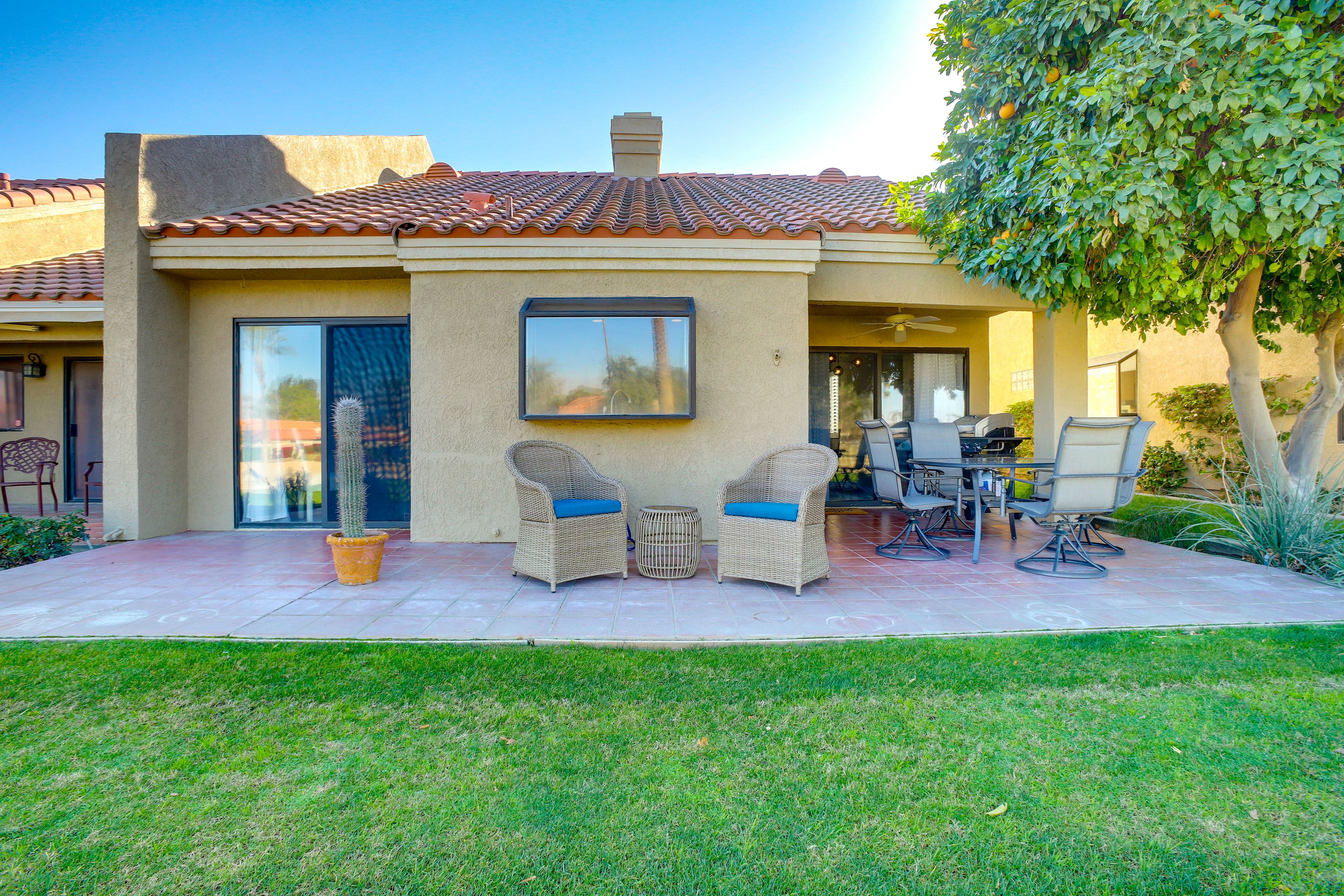 A house with a patio, two wicker chairs with cushions, a table, and a cactus.
