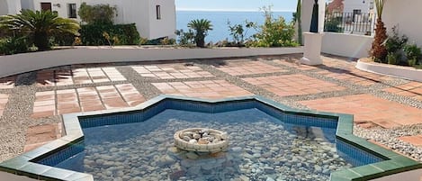 A swimming pool with a circular stone feature in the center, surrounded by a white wall and a tiled deck.