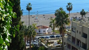 Blick auf einen Strand mit Palmen, ein weißes Gebäude mit rotem Dach und ein Parkplatz mit Autos.