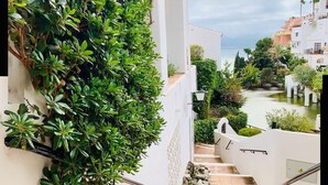 Eine Treppe mit einer üppigen, grünen Hecke an einer Seite, die zu einem Innenhof mit Blick auf das Meer und andere Gebäude führt.