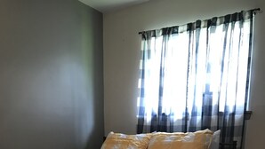 Een bed met een geruit sprei, een houten nachtkastje en een raam met dunne gordijnen.