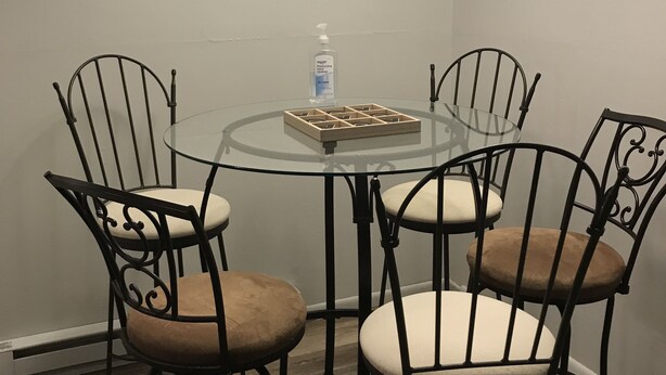 Een eetgedeelte met een ronde glazen tafel, omringd door vier metalen stoelen met zachte zittingen. Boven de tafel hangt een ronde wandklok met cijfers.