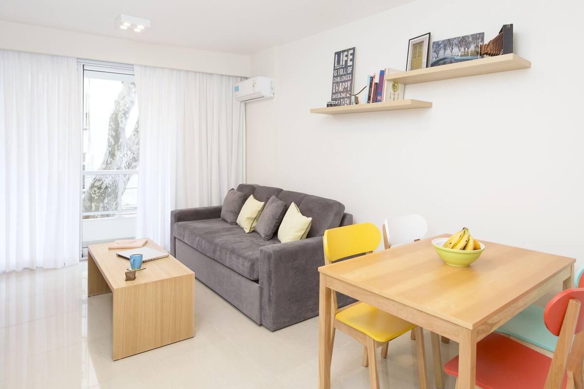 Un salon moderne avec un canapé gris, une table basse en bois, une table à manger avec des chaises et une petite bibliothèque.