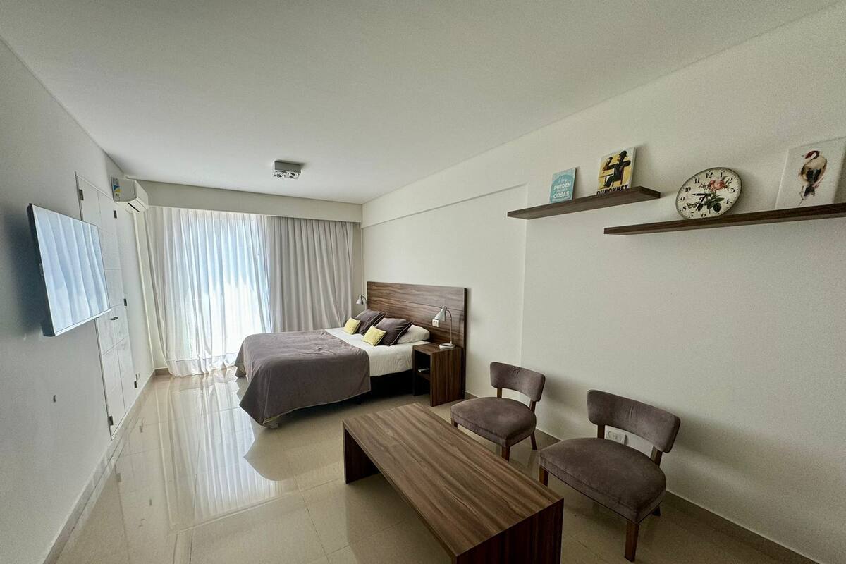 Une chambre d’hôtel moderne équipée d’un lit, d’un bureau, de deux chaises, d’une télévision et d’une étagère ornée d’objets décoratifs.
