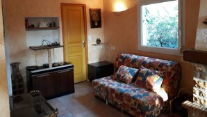 A cozy living room with a sofa, a wooden door, a window, and a kitchen area with cabinets and a sink. - Holiday Home 'Frejus' with Shared Pool, Shared Terrace and Air Conditioning (Frejus)