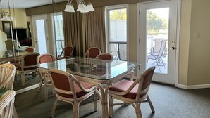 Een eetgedeelte met een glazen tafel, omringd door rieten stoelen, een kroonluchter en een uitzicht door Franse deuren op een terras.