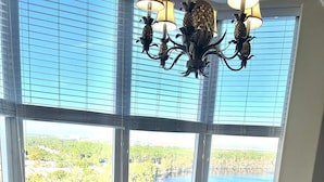 Un comedor con mesa de vidrio y sillas de madera, ventanas grandes con persianas y vista a un lago y bosque.