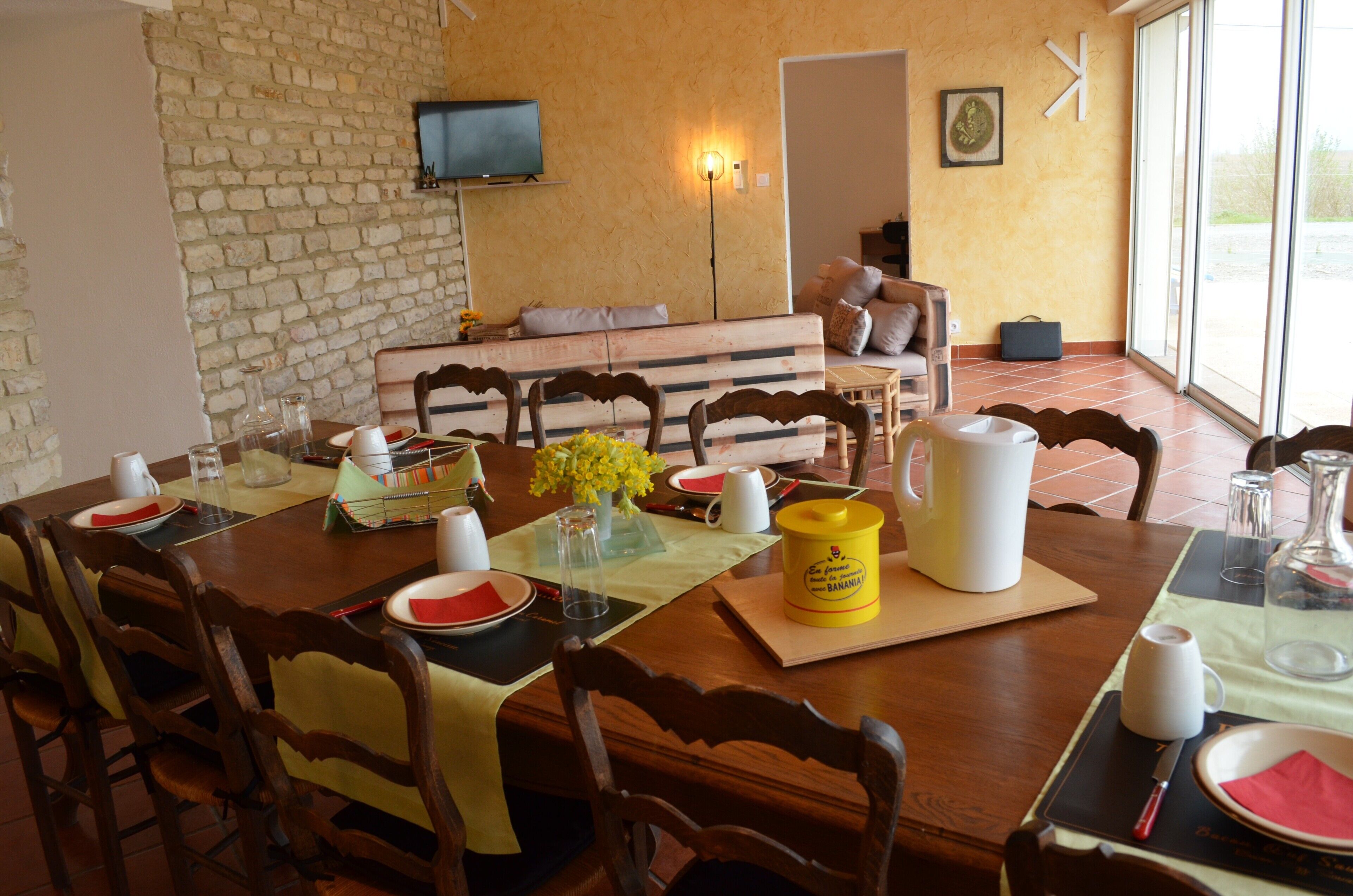 Un coin repas avec une table en bois dressée pour quatre personnes, un banc à l’arrière-plan et une grande fenêtre donnant sur un beau paysage.
