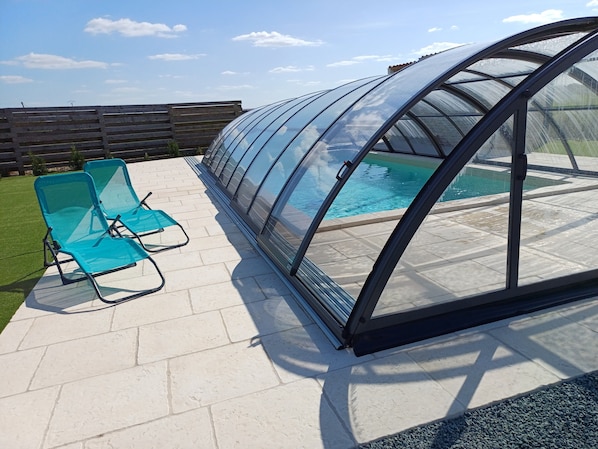 Una estructura tipo invernadero con una piscina, dos sillones azules y una valla de madera al fondo.