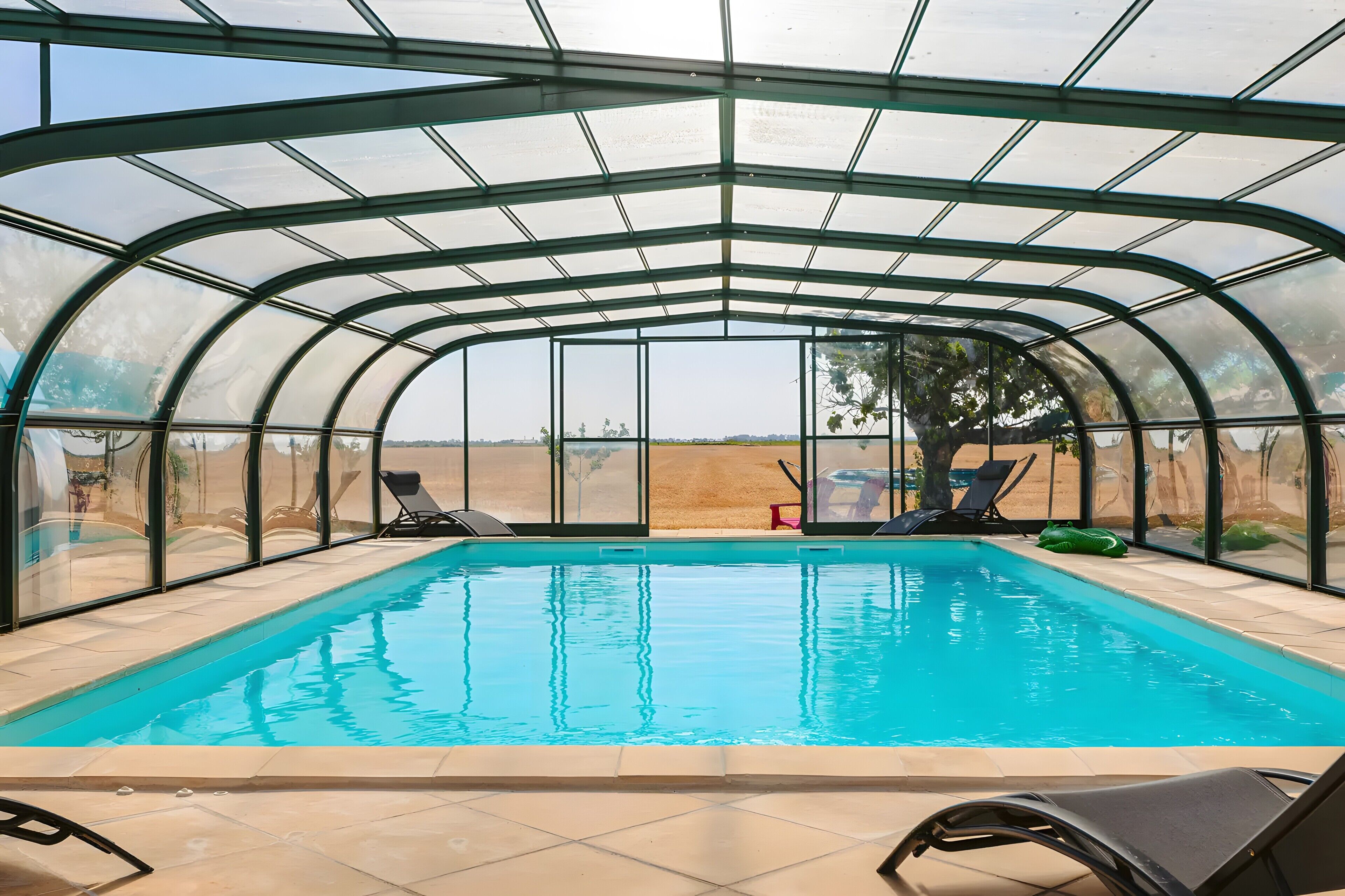 A covered swimming pool with clear glass walls, lounge chairs, and a view of the landscape.