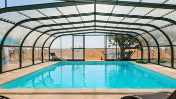 A covered swimming pool with clear glass walls, lounge chairs, and a view of the landscape.