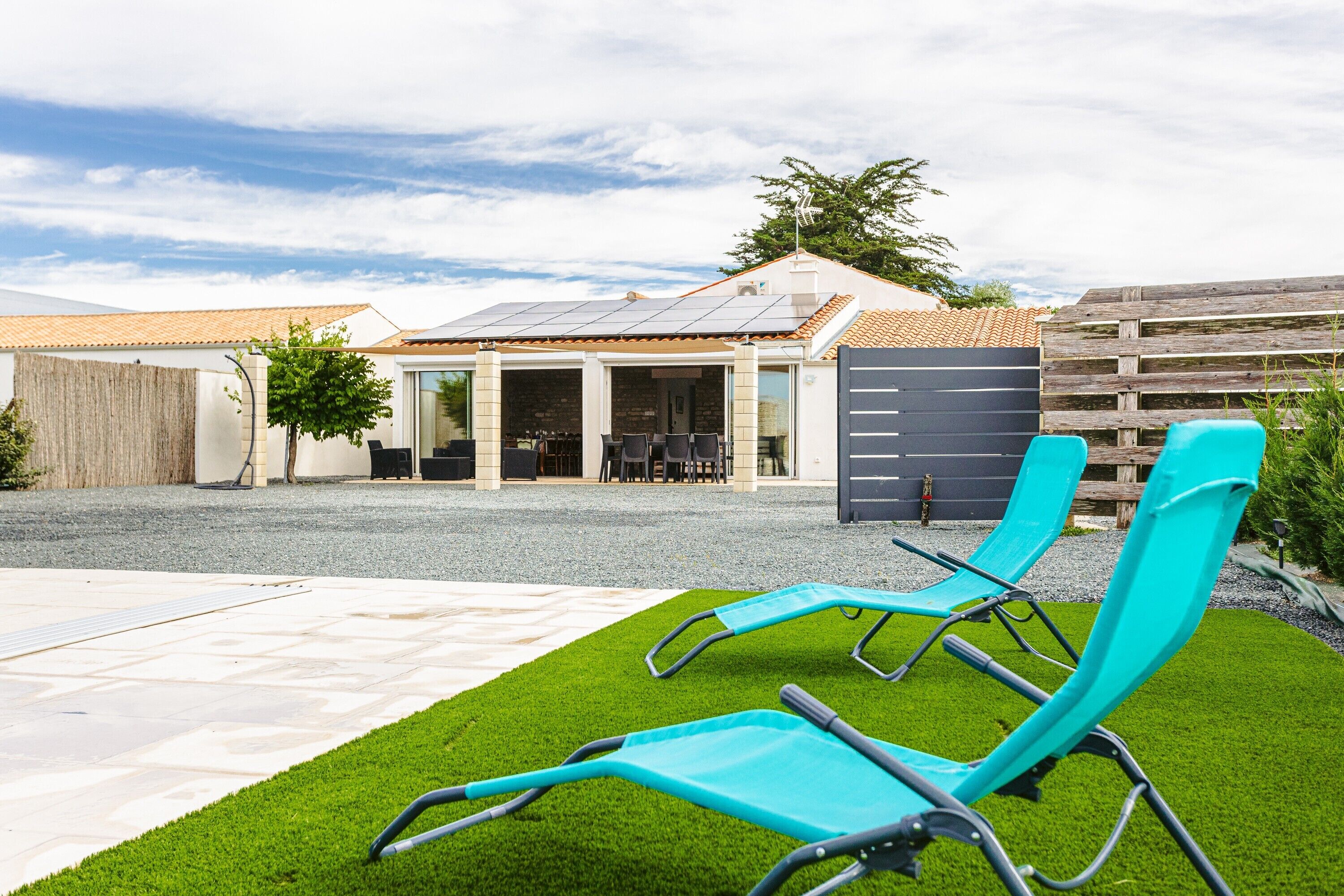 Une terrasse avec deux fauteuils de jardin bleus, un espace extérieur aménagé et couvert, et un bâtiment doté d’un toit en tuiles et d’une clôture en bois.