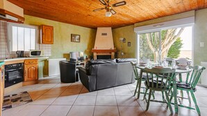 A kitchen with a microwave, oven, and wooden cabinets. There is a black leather sofa and a dining table with chairs. A ceiling fan is visible. - Holiday Home 'Gîte Le Petit Sableau' with Shared Pool & Spa, Private Terrace, Wi-Fi (MARANS)
