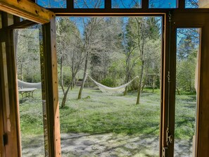 Vista a través de una ventana que muestra una habitación con una hamaca, una mesa y un sendero que lleva a una escalera en un área boscosa.