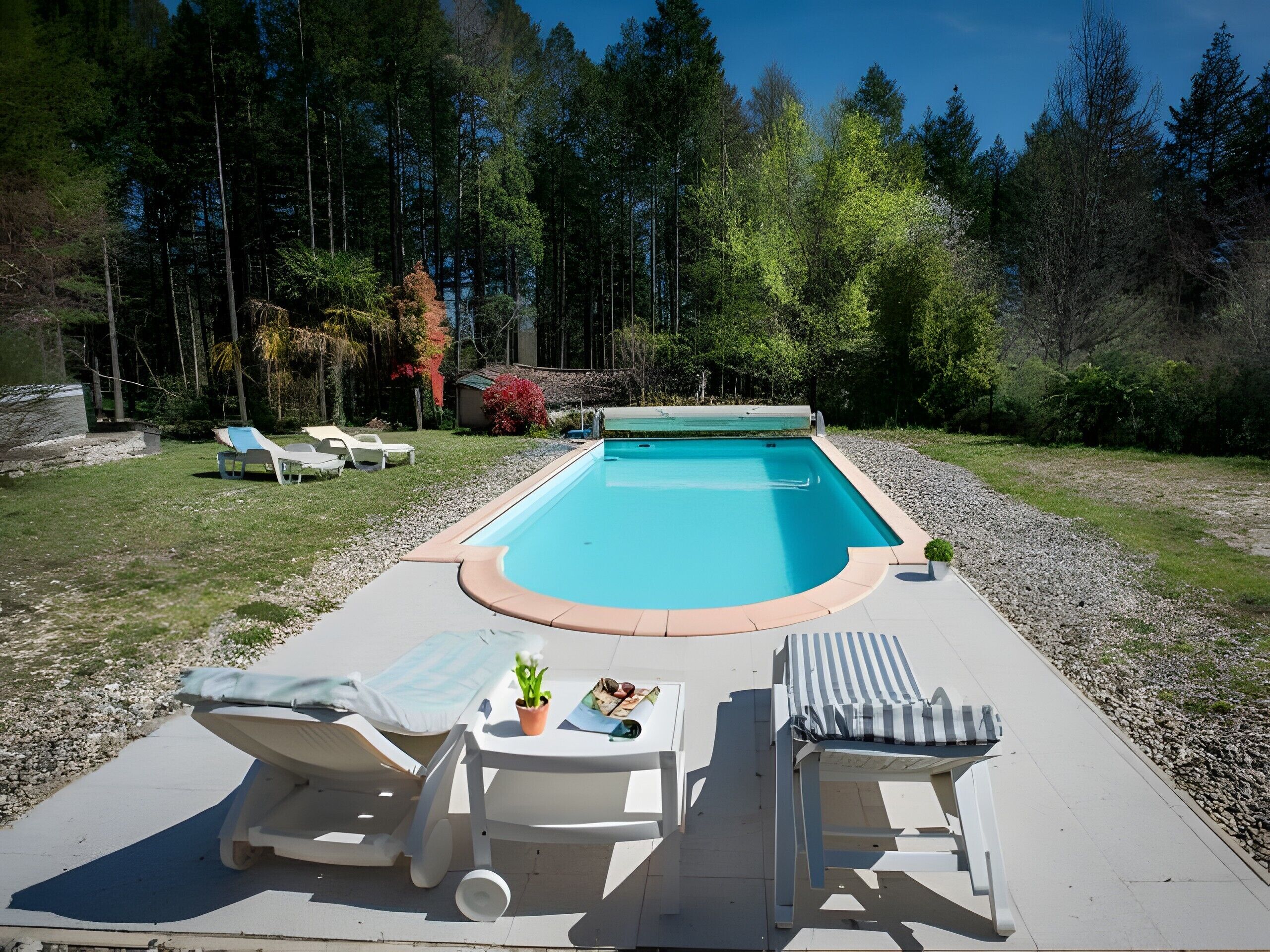 Una piscina rectangular con borde curvo, rodeada por un camino de piedrecitas. Hay sillas de descanso y una pequeña mesa con una planta y gafas de sol.