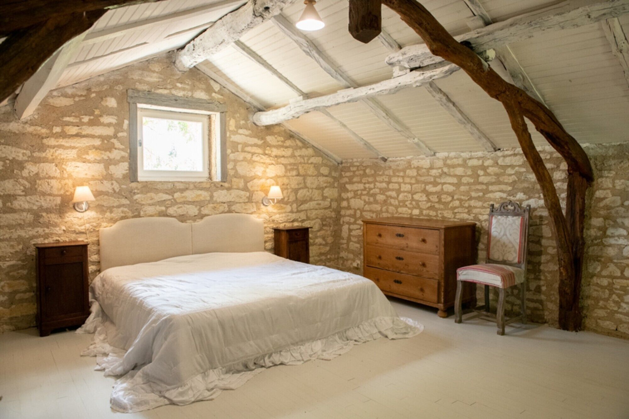 A bedroom with a stone wall, a bed, a dresser, a chair, and two wall-mounted lamps.