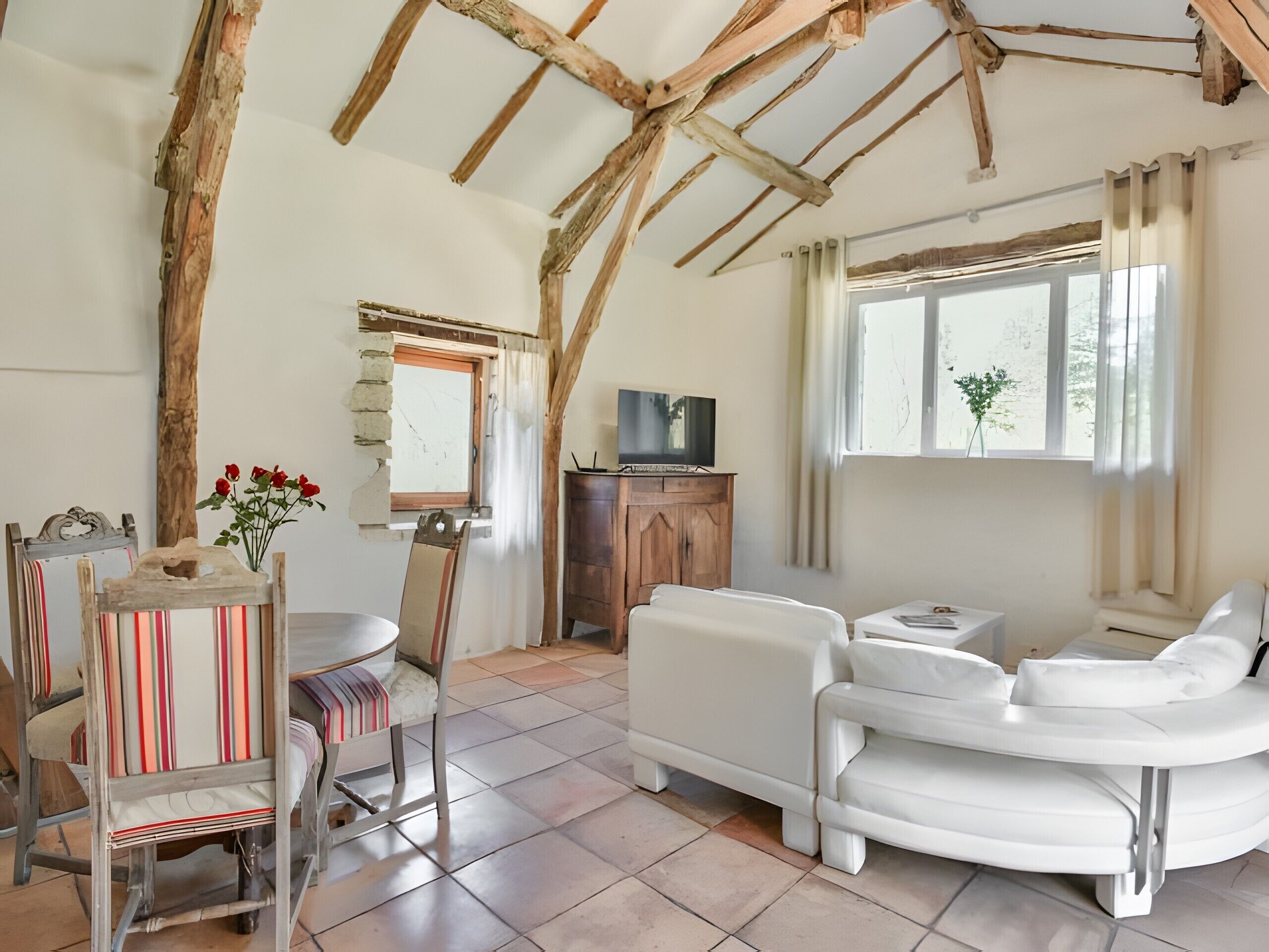 A rustic interior with exposed wooden beams, a white sofa, a dining table with chairs, and a television on a wooden cabinet.