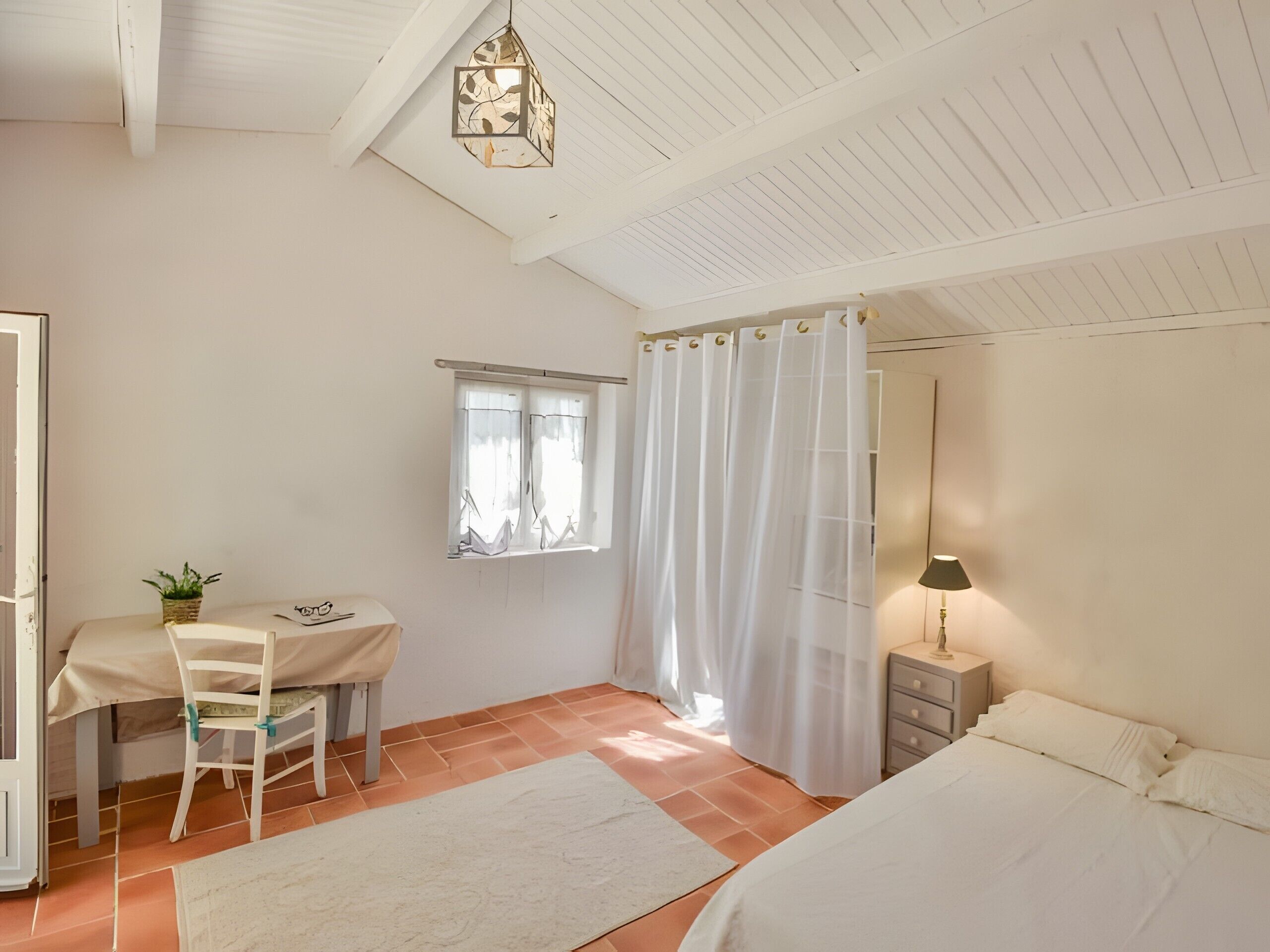A bedroom with a bed, a desk with a chair, a window with curtains, a nightstand with a lamp, and a hanging light fixture.