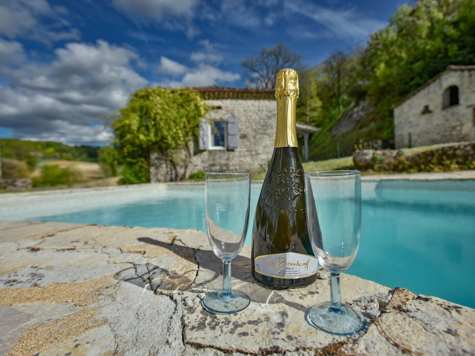Eine Champagnerflasche und zwei Gläser auf einer Steinoberfläche an einem Pool.