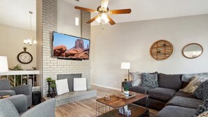 A living room with a sectional sofa, a coffee table, a TV mounted on a brick wall, a ceiling fan, and a fireplace. - 4 Bed Shuffleboard& Private Pool Retreat in Peoria (Peoria)