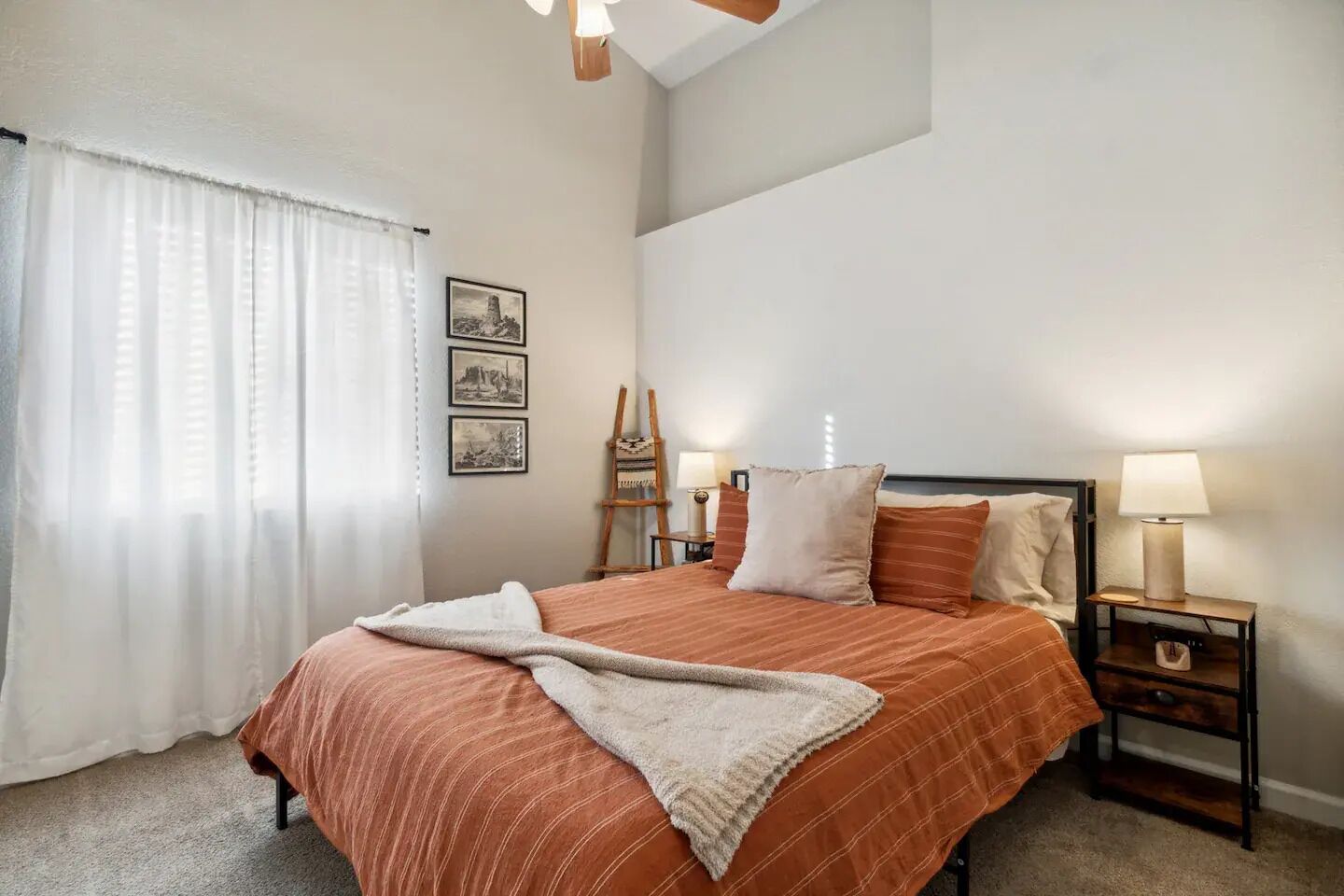 A bedroom with a bed, two bedside tables with lamps, a ladder, and three framed pictures on the wall.