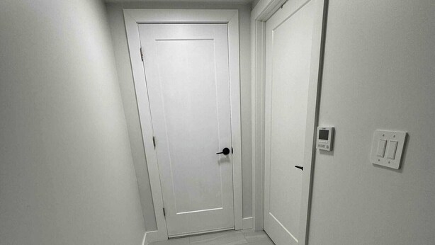 Un pasillo estrecho con una puerta blanca al final, un interruptor de luz y un termostato en la pared.