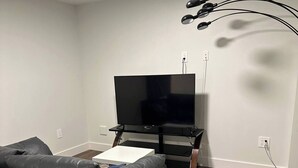Una sala de estar con un sofá de cuero negro, un televisor de pantalla plana sobre un mueble, una mesita blanca pequeña y una alfombra estampada sobre piso de madera.