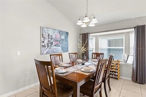 A dining area with a wooden table set for six, surrounded by chairs. There is a chandelier above the table, and a window with blinds to the right.