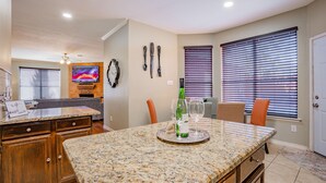 Una cocina con encimeras de granito, armarios de madera, una zona de comedor con una mesa y sillas, y un televisor montado en la pared.
