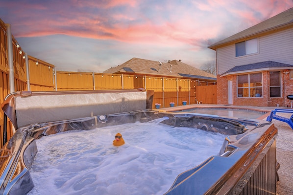 A hot tub with a person in it, surrounded by a wooden fence and a house in the background.