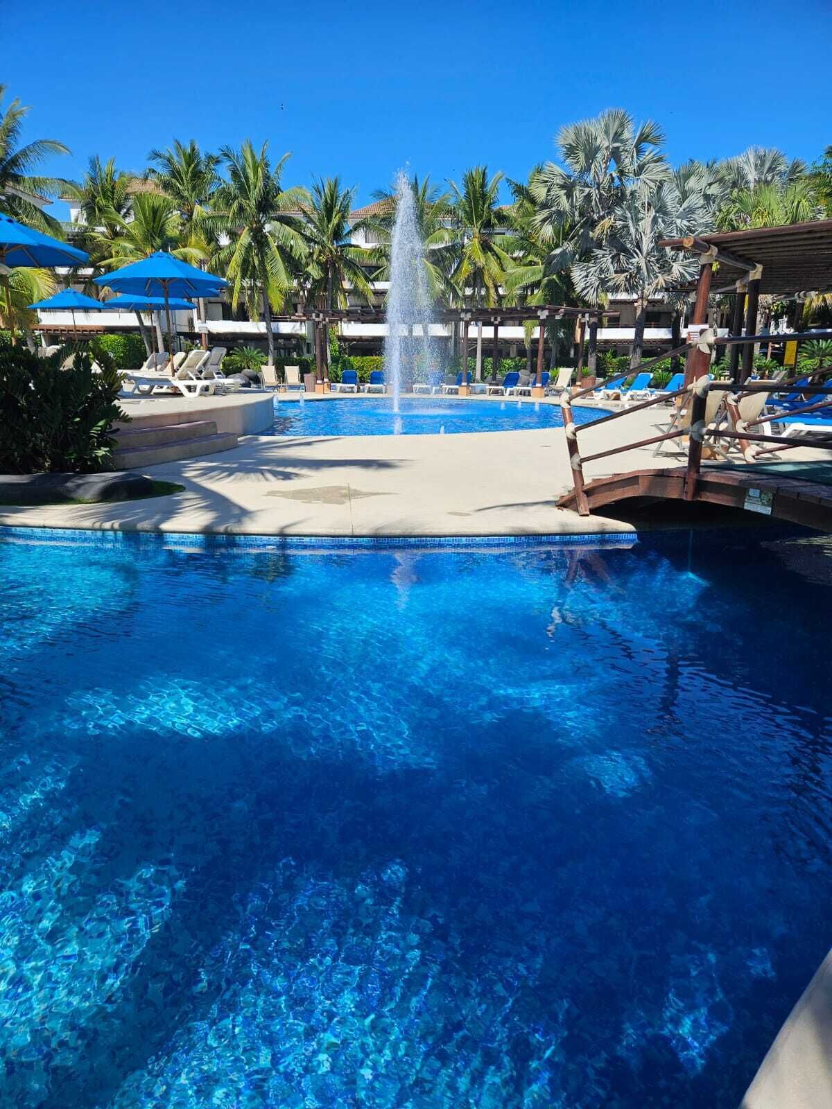 A clear blue pool with a central fountain, surrounded by lounge chairs and umbrellas, with palm trees in the background.