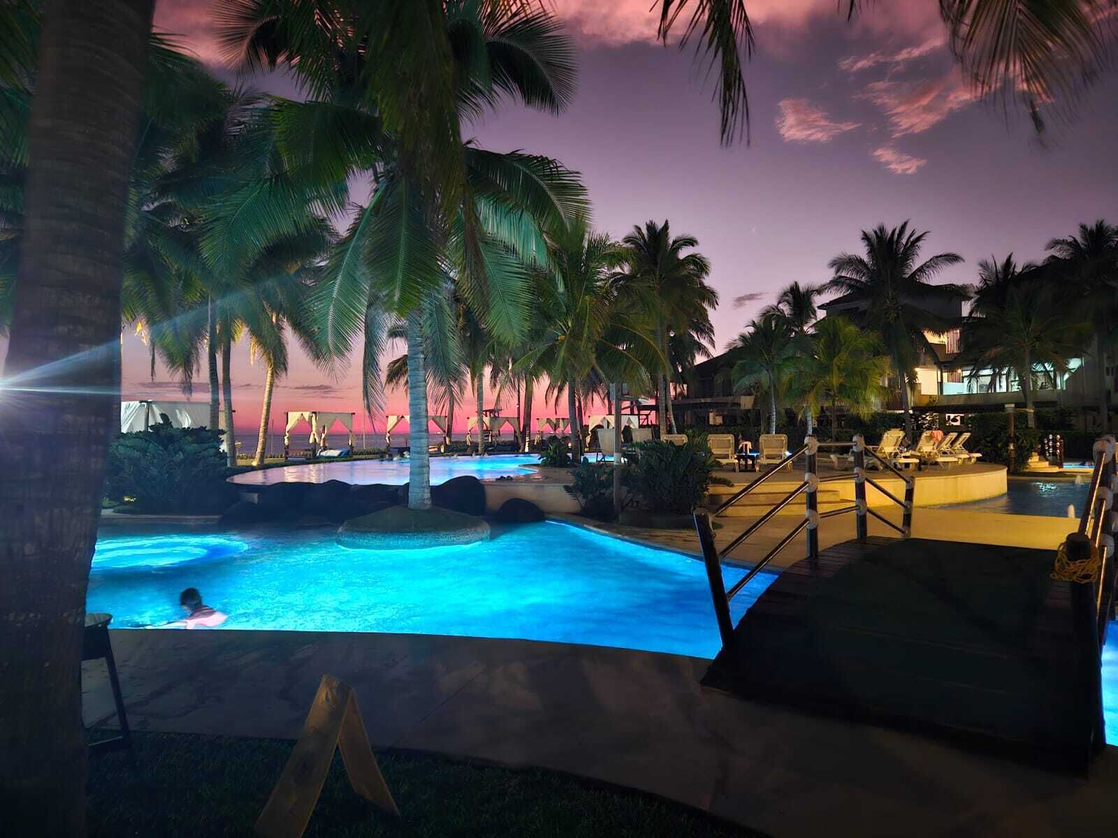 Zona de piscina con palmeras y sillas de descanso, rodeada por un edificio con un cielo rosado al fondo.