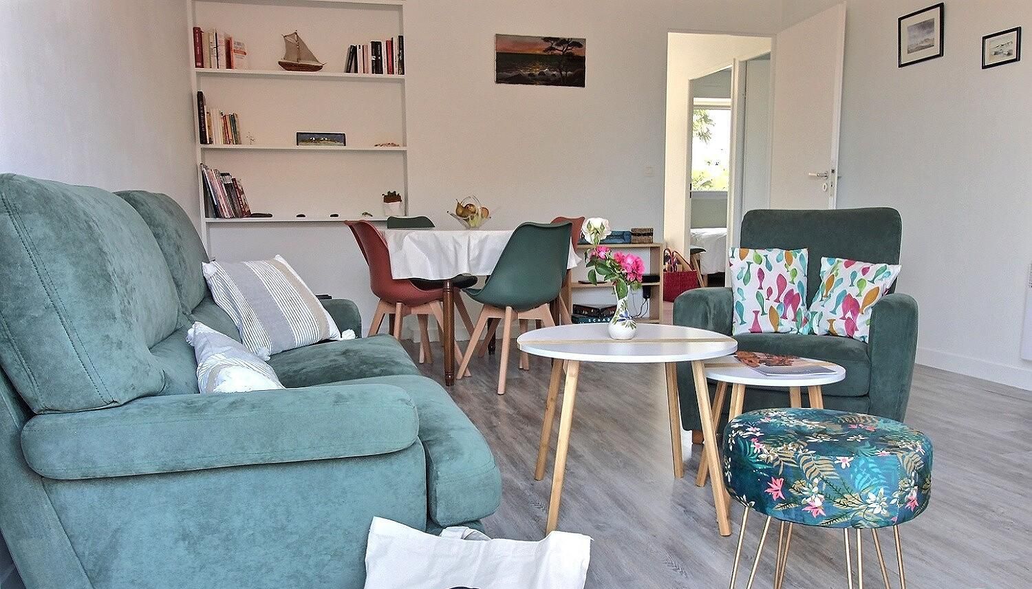 Un salon avec un canapé vert, une table basse ronde et un pouf à motifs floraux. On y trouve une bibliothèque remplie de livres et d’objets décoratifs, ainsi qu’un coin repas avec une table et des chaises.