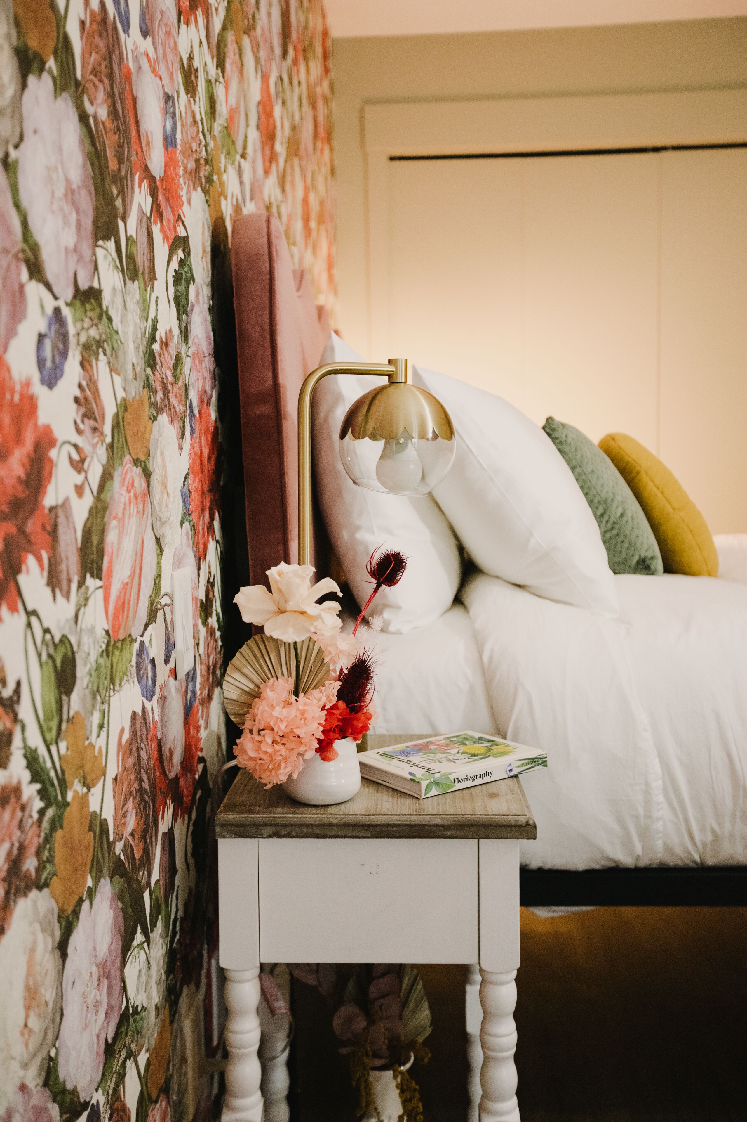 Un lit bien fait, avec une literie blanche, une petite table de chevet avec une lampe et un papier peint à motifs floraux.